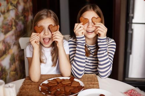 Charmante, lustige und lachende Mädchen halten herzförmige Kekse in der Hand, schließen die Augen und albern herum. Valentinstag und Frauentag niedlich Küche