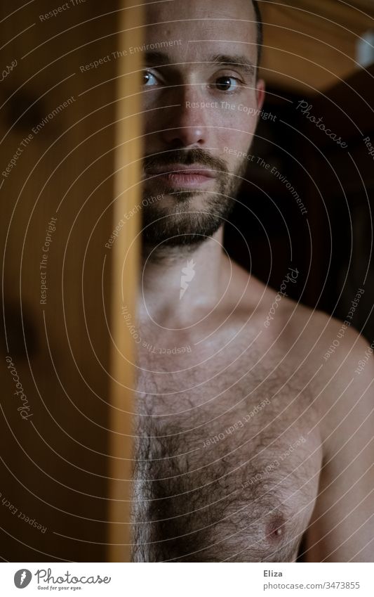 Ein Mann mit nacktem Oberkörper, der hinter einer Holztür hervor direkt in die Kamera blickt Oberkörperfrei Brust in die Kamera schauen Blick in die Kamera