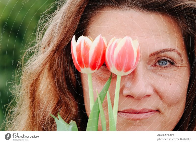 Porträt einer schönen Frau, die sich über den Frühling freut, mit zwei Tulpen vor ihrem rechten Auge, lächeln Freude Gesicht Mensch Glück hübsch Kaukasier