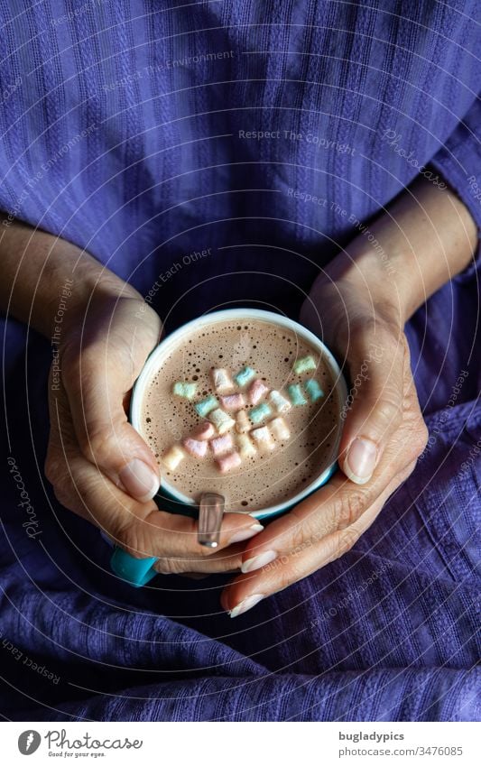 Eine Frau in einem lila Kleid hält mit beiden Händen eine türkise Tasse mit Cappuccino der mit Minimarshmallows dekoriert ist Marshmallows Tasse festhalten