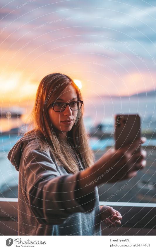 Junge Frau beim Egoismus mit dem Ozean im Hintergrund Selfie Selbst Smartphone Foto Fotografieren Sonnenuntergang Balkon Quarantäne grau Sweatshirt blond Brille