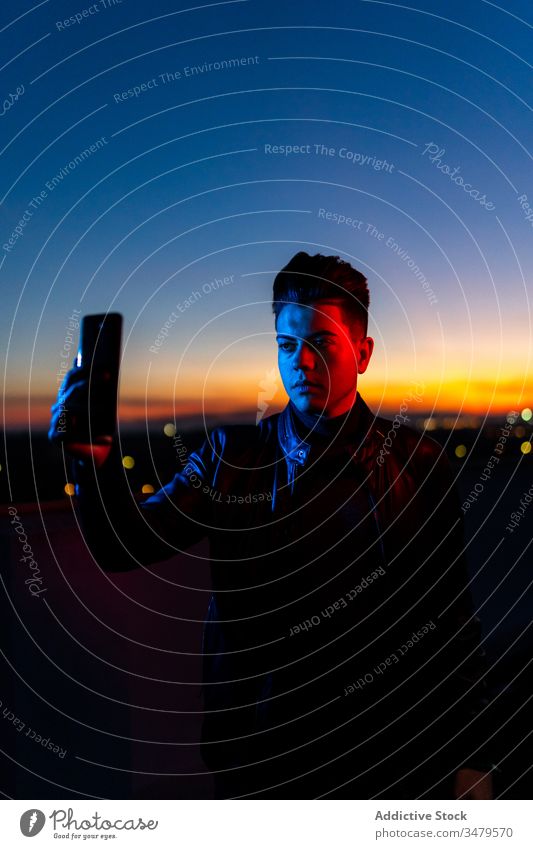 Junger Mann nimmt sich abends selbst die Zeit Smartphone Selfie neonfarbig Handy Sonnenuntergang dunkel blau Licht Himmel Stil ernst Farbe Apparatur Gerät