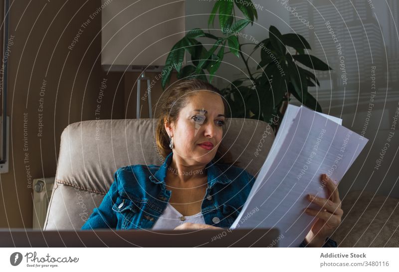 Erwachsener Freiberufler mit Laptop liest Dokument Frau freiberuflich lesen Papier benutzend heimwärts reif Fenster Armsessel sitzen Gerät Apparatur Drahtlos
