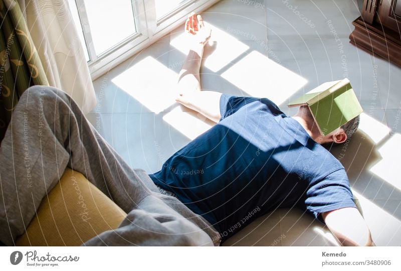 Auf dem Boden liegender junger Mann mit einem Buch im Gesicht, während er die durch das Fenster einfallende Sonne genießt. Konzept von Zuhause bleiben, Freiheit, Langeweile...