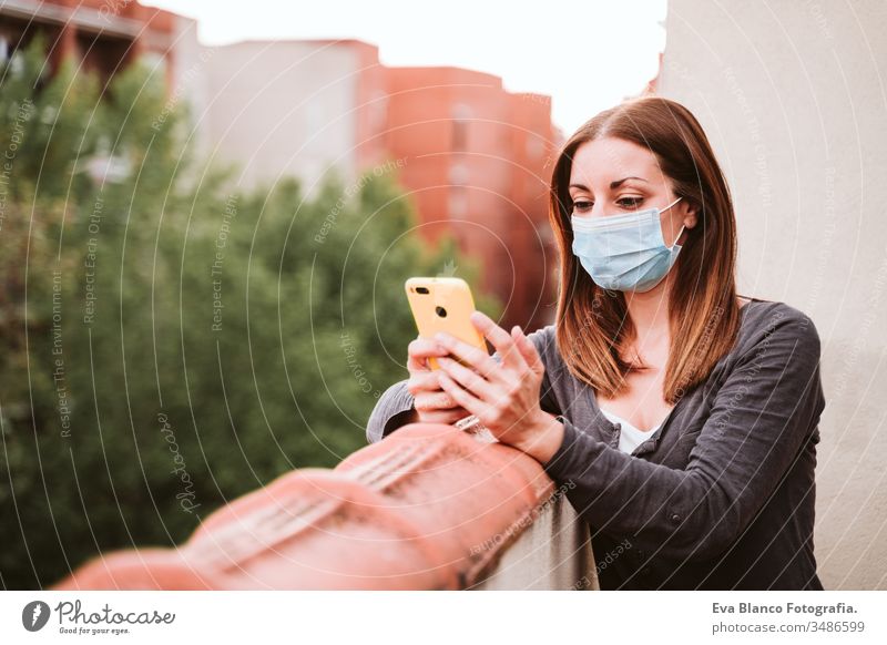 junge Frau zu Hause auf einer Terrasse, die eine Schutzmaske trägt, ein Mobiltelefon benutzt und einen sonnigen Tag genießt. Coronavirus Covid-19-Konzept Handy
