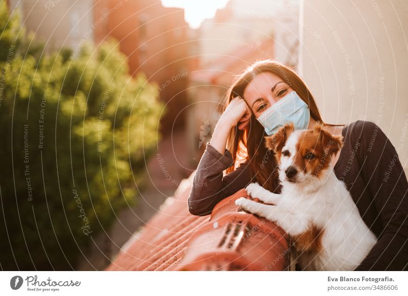 junge Frau zu Hause auf einer Terrasse mit Schutzmaske. Sie umarmt ihren süßen Hund Jack Russell. Coronavirus Covid-19-Konzept Haustier covid-19 schützend