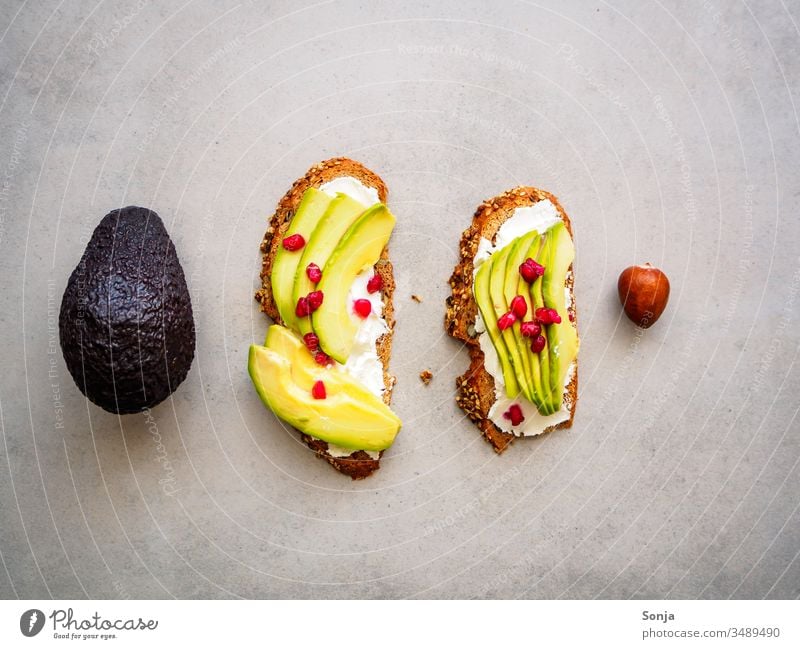 Vollkornbrot mit Avocado und Granatapfelkernen, eine ganze Avocade und ein Kern auf einem grauen Hintergrund, Draufsicht, gesundes Frühstück vollkornbrot