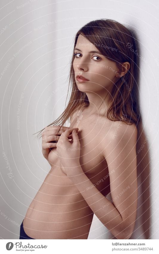 Sexy, oben ohne, schlanke, junge, brünette Frau in Dessous, die mit ihren Händen an einer Innenwand steht und ihre Brüste bedeckt, was der Kamera einen schwülen Blick verleiht