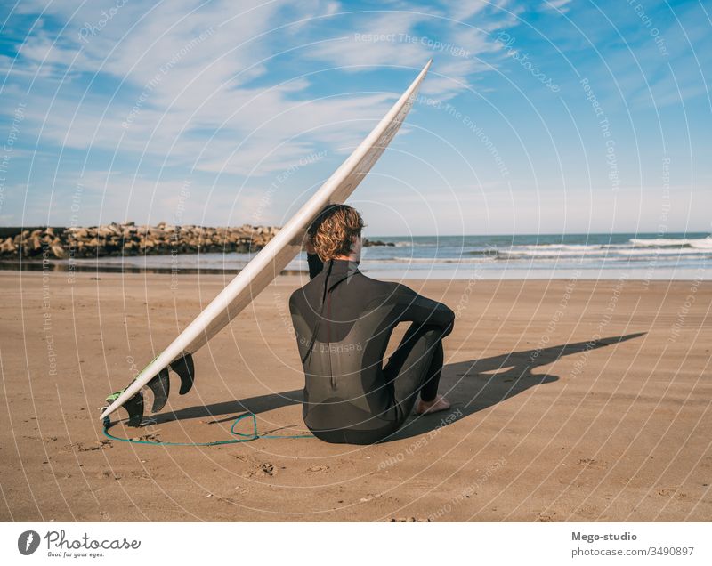 Surfer, der am Sandstrand und neben dem Surfbrett sitzt. Mann Brandung Wasser Sport Surfen MEER Meer im Freien sportlich Küstenlinie Wellen Hintergrund