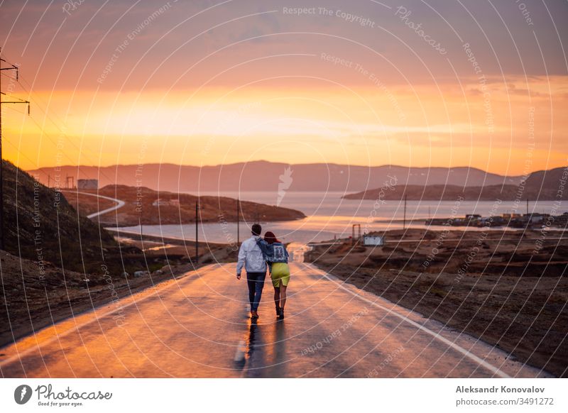 Junges Paar auf dem Weg zur Barentssee auf einer nassen Straße in einem Sonnenuntergang Nasse Straße Golf Meer Tundra polar Dorf wählen Strommast Asphalt