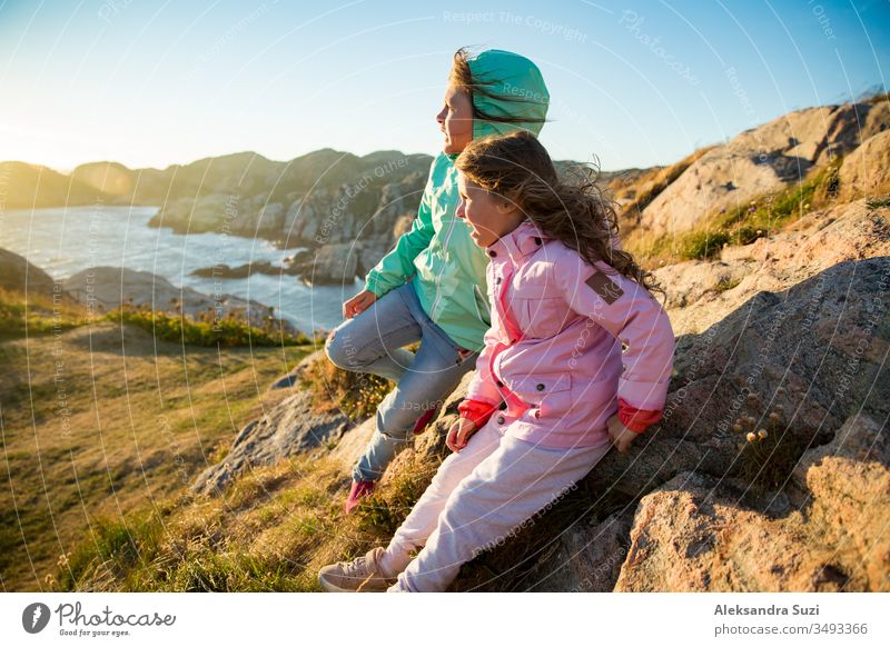 Zwei kleine Mädchen spielen an der felsigen Nordküste. Sitzen, lachen, umarmen, erkunden die Küstenfelsen. Reisen und ein großes Abenteuer in Norwegen erleben. Wunderschöne Aussicht auf Fjord und Berge bei Sonnenuntergang.