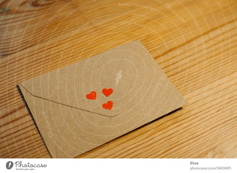 Ein Briefumschlag mit drei kleinen roten Herzen auf einem braunen Holztisch; Konzept Liebe, Liebesbrief Grüße verliebt Post Farbfoto Kommunizieren schreiben