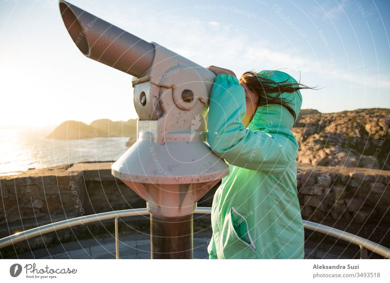 Ein junges Mädchen steht am felsigen nördlichen Meeresufer, schaut durch ein touristisches Fernrohr und erkundet die Küstenfelsen. Reisen und Abenteuer in Norwegen erleben. Wunderschöne Aussicht auf Fjord und Berge bei Sonnenuntergang
