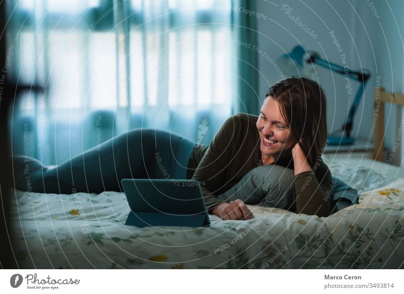 Lächelnde junge Frau liegt auf dem Bett, während sie mit ihrer Familie einen Videoanruf macht. Attraktive Frau, die ihr digitales Tablett während der Quarantäne zu Hause benutzt.