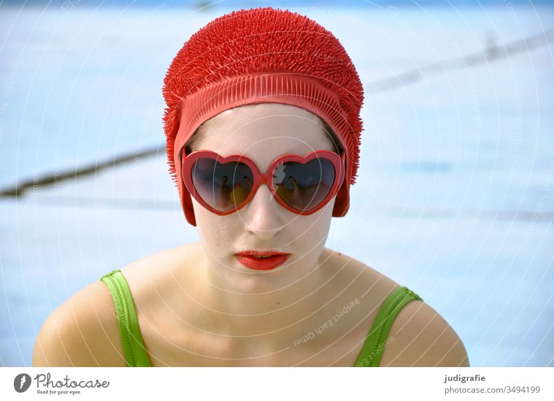 Das Mädchen mit der schönen roten Badekappe und dem grünem Badeanzug schaut durch eine Herzchen-Sonnenbrille direkt in die Kamera. Eine Sommerliebe. Frau