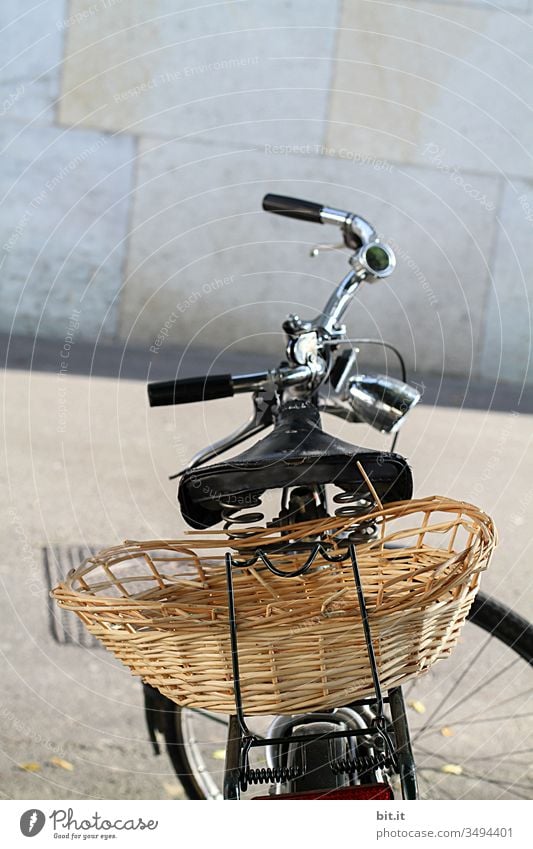 Fahrrad mit Lenker, Sattel und Korb zum Einkaufen und Transport auf dem Gepäckträger steht geparkt auf der Straße, in der Stadt, vor grauer Wand im Hintergrund.