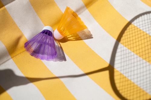 Zwei bunte Federbälle mit dem Schatten eines Federballschlägers auf einem gelb weiß gestreiftem Hintergrund sommerlich Sport Hobby spielen Badminton