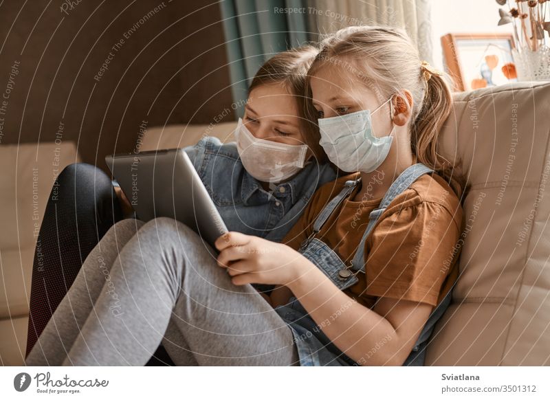 Zwei süße Mädchen mit medizinischen Masken sitzen mit einer Tablette auf dem Sofa. Schulmädchen machen ihre Hausaufgaben. Fernunterricht während der COVID-19-Quarantäne.