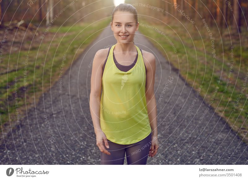 Lächelnde, sportlich-sportliche junge Frau, die auf einer geteerten Bahn durch Wälder, die vom warmen Schein der Sonne beleuchtet werden, in einem gesunden, aktiven Lebensstil trainiert