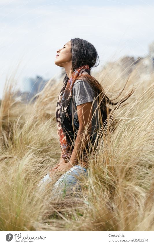 Porträt einer jungen brünetten Frau mit langen Haaren, die einen Schal um den Hals trägt und mit ruhigem Gesichtsausdruck auf den Knien in einem Weizenfeld sitzt