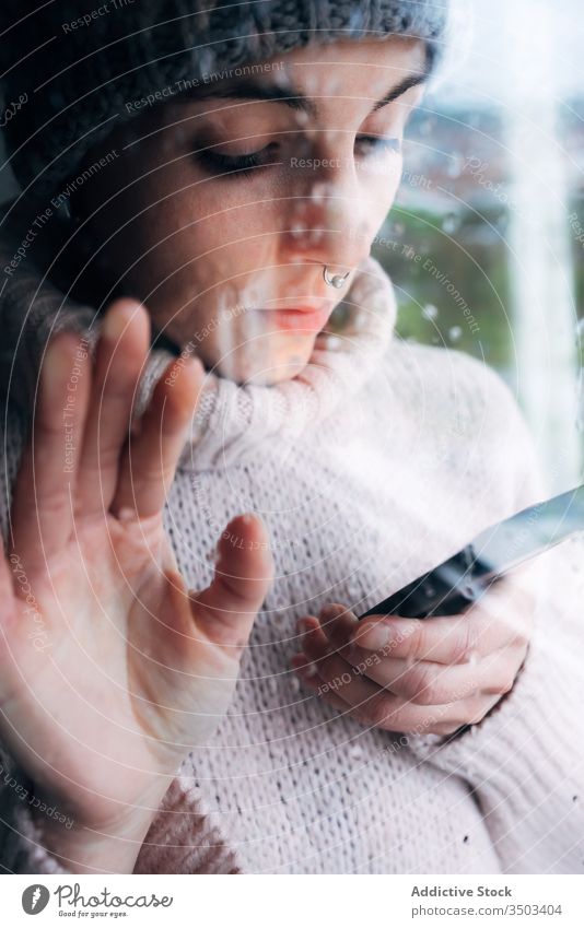 Junge Frau mit Smartphone am Fenster stehend heimwärts benutzend einsam traurig Selbstisolierung Isolation Browsen Apparatur Coronavirus covid-19 Quarantäne