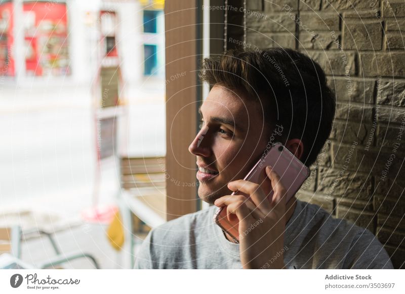 Fröhlicher junger Mann spricht auf Smartphone in der Nähe des Café-Fensters sprechen Lächeln modern Glück Anruf männlich fettarm Restaurant Telefon Mobile