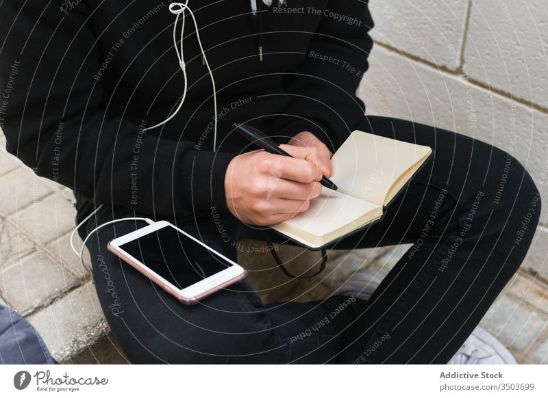 Crop-Schüler, die Musik hören und in einem Notizbuch schreiben Mann Straße Notebook zuhören Smartphone lässig Schritt männlich Großstadt Notizblock Hinweis