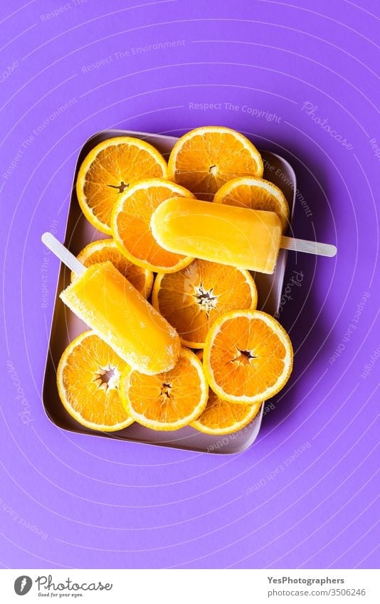Eis am Stiel in Orange auf violettem Hintergrund obere Ansicht Kindheit Zitrusfrüchte klassisch kalt farbenfroh Farben Kontrast ausschneiden lecker Dessert