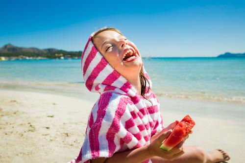 Süßes fröhliches kleines Mädchen isst Wassermelone und lacht am Sandstrand. Türkisfarbenes Meer, blauer Himmel, sonniger Sommertag. Mallorca, Spanien Aktivität