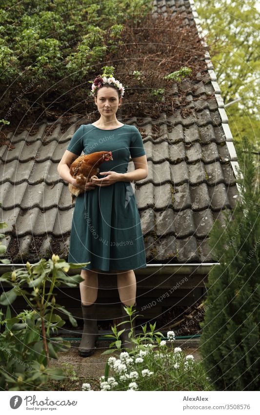 Junge Frau mit Blumenkranz im Haar steht vor Gartenhaus und hält braunes Huhn im Arm Zentralperspektive Schwache Tiefenschärfe Tag Außenaufnahme Farbfoto Idylle