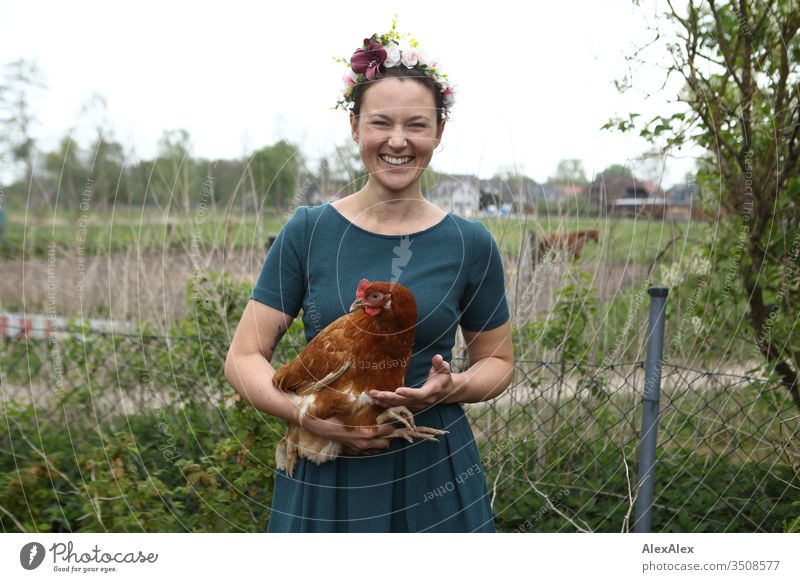 Junge Frau mit Blumenkranz im Haar steht im Garten hält braunes Huhn im Arm Zentralperspektive Schwache Tiefenschärfe Tag Außenaufnahme Farbfoto Idylle feminin