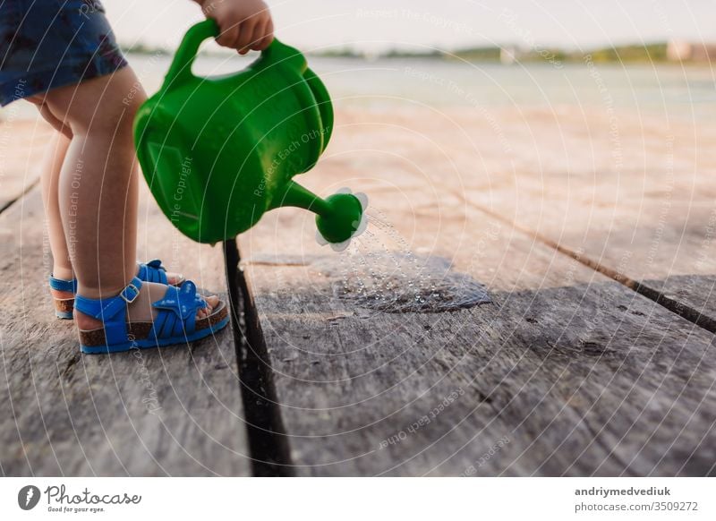 Ein kleines Mädchen spielt mit der Gießkanne einer Holzbrücke. Frühling und Sommer. Gartenarbeit. grüne Gießkanne Bewässerung Dose Wasser Baby wenig Kind