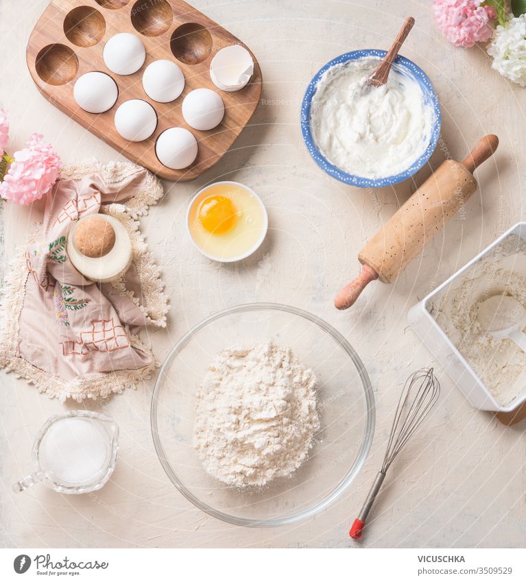 Moderner Backhintergrund mit Mehl in Schüssel, Eier und Zutaten auf weißem Küchentischhintergrund, Draufsicht. Hausgemachtes Backen. Rezepte für Kekse, Kuchen und Torten