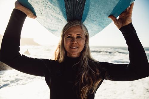 Lächelnde ältere Frau trägt Surfbrett am Strand und schaut in die Kamera Senior Frauen Urlaub Surfen Erwachsener graue Haare Lifestyle Freude Kaukasier Sport