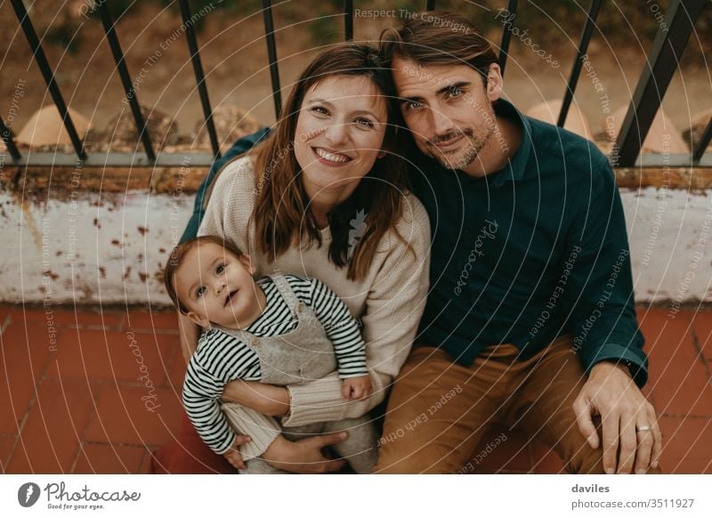 Wunderschönes Porträt eines gut aussehenden Mannes und einer schönen Frau mit ihrem kleinen Sohn, die in die Kamera schauen, während sie auf dem Boden ihrer heimischen Terrasse sitzen.
