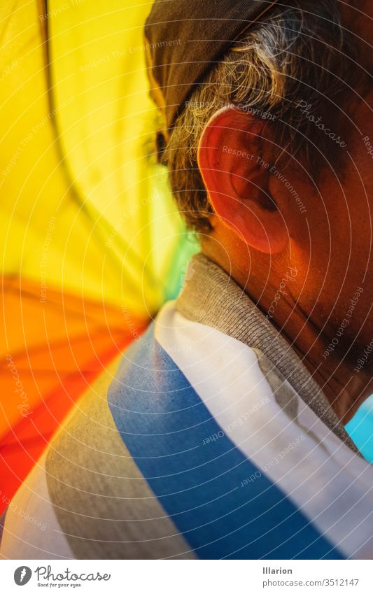 Nahaufnahme des Ohrs eines alten Mannes mit einem Regenbogenschirm im Vordergrund alter Mann Holunderbusch regenbogenfarben Regenbogentuch Farbe Farben