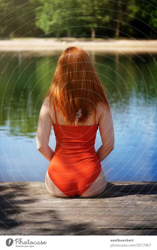 Rückansicht einer Frau im roten Badeanzug auf einem Holzsteg sitzend Badesee See Baden schwimmen Schwimmsport Rückseite Rücken hinten unkenntlich Person