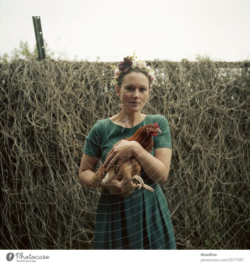 Junge Frau mit Blumenkranz im Haar steht im Garten hält braunes Huhn im Arm Zentralperspektive Schwache Tiefenschärfe Tag Außenaufnahme Farbfoto Idylle feminin