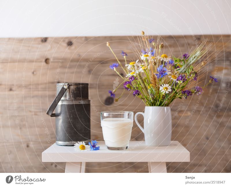 Ein Glas frische Milch, ein Strauß Wiesenblumen in einer Vase und eine alte Milchkanne auf einem weißen Hocker. Ländliches Motiv trinkglas Blumen rustikal weiss