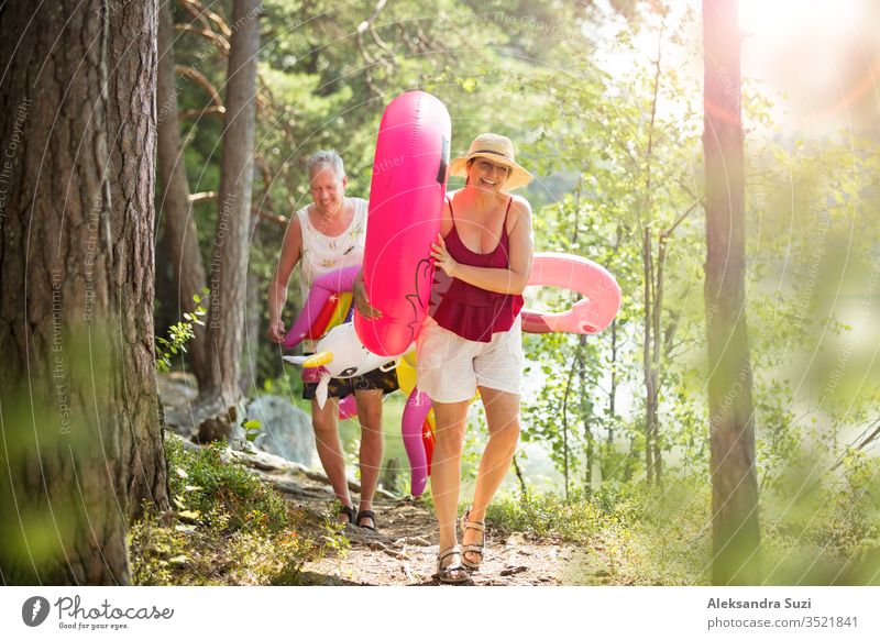 Ein älteres Ehepaar, das auf einem Waldweg am Meeresufer spazieren geht und einen aufblasbaren Riesenflamingo und ein Einhorn in der Hand hält. Lustige aktive Rentner genießen Sommerurlaub am Strand in Nordeuropa