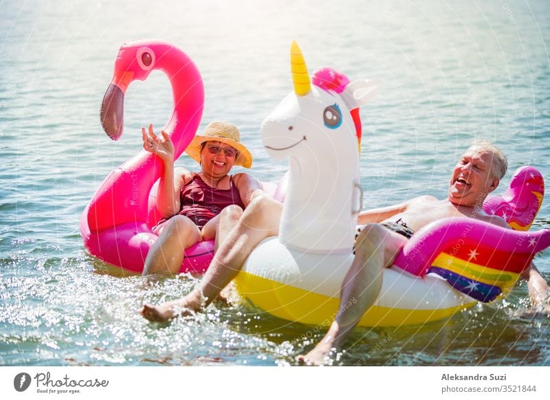 Älteres Paar amüsiert sich auf aufblasbaren Flamingos und Einhörnern. Lustige aktive Rentner, die zusammen glücklich den Sommerurlaub am Strand in Europa genießen, lachen, den Narren spielen, Wasser spritzen.