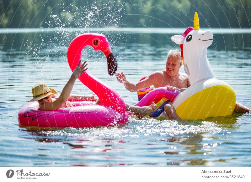 Älteres Paar amüsiert sich auf aufblasbaren Flamingos und Einhörnern. Lustige aktive Rentner, die zusammen glücklich den Sommerurlaub am Strand in Europa genießen, lachen, den Narren spielen, Wasser spritzen.
