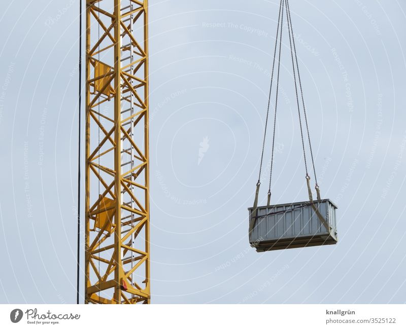 Gelber Baukran der am Ausleger einen mittelgroßen grauen Metallcontainer an Ketten hängen hat Kran Container Baustelle hoch Schweben Sicherheit bauen