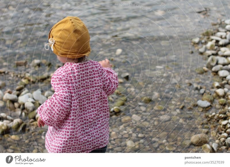 Weibliches Kleinkind spielt am flußbett mit Steinen spielen wasser isar steine werfen kleidung sonnenschutz gelb blumenmuster frühling sommer Außenaufnahme Tag