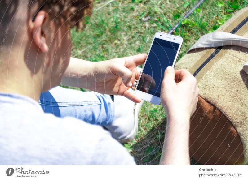 Mann, der ein Mobiltelefon benutzt und mit einer Sporttasche auf dem Rasen sitzt Bildschirm Reflexion & Spiegelung Gerät Rücken Detailaufnahme Zoom Texten