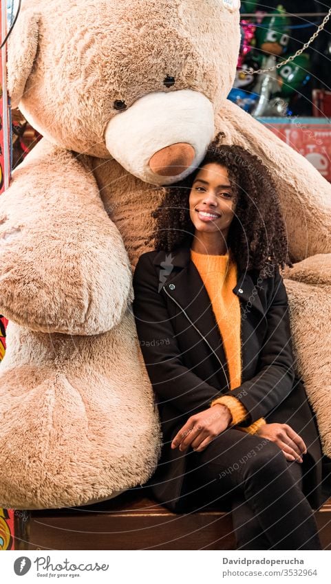 Glückliche ethnische Frau sitzt mit riesigen Teddybär Bär positiv heiter Lächeln Spielzeug Plüsch groß Afroamerikaner Mantel jung Lifestyle sitzen Freude