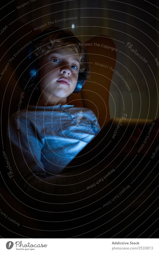 Vertikales Bild eines sitzenden Kindes, das nachts mit abgelenktem Blick auf ein Tablett mit Kopfhörern schaut Junge Abgelenkt Vorstellungskraft Kino Multimedia