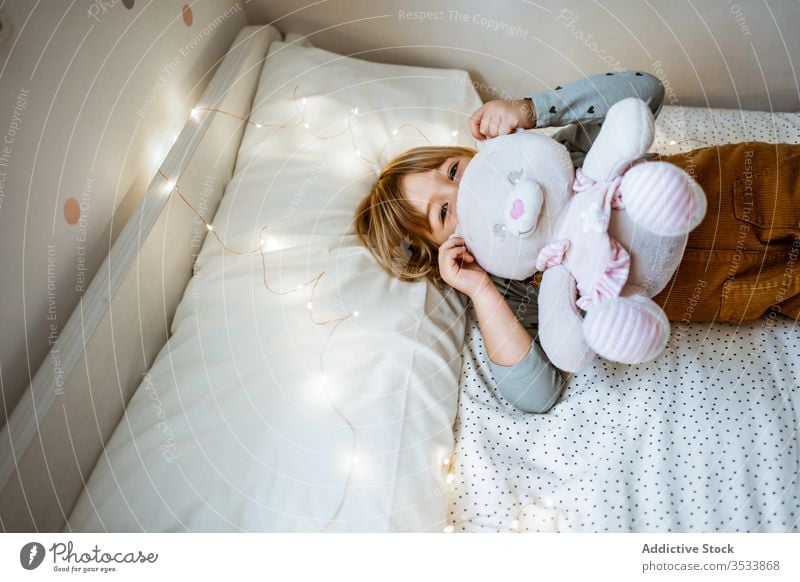 Glückliches Mädchen spielt auf weichem Bett spielen Spielzeug Umarmung Lachen Lichterkette Plüsch Lügen aufgeregt Kind Liebe Girlande Schlafzimmer wenig