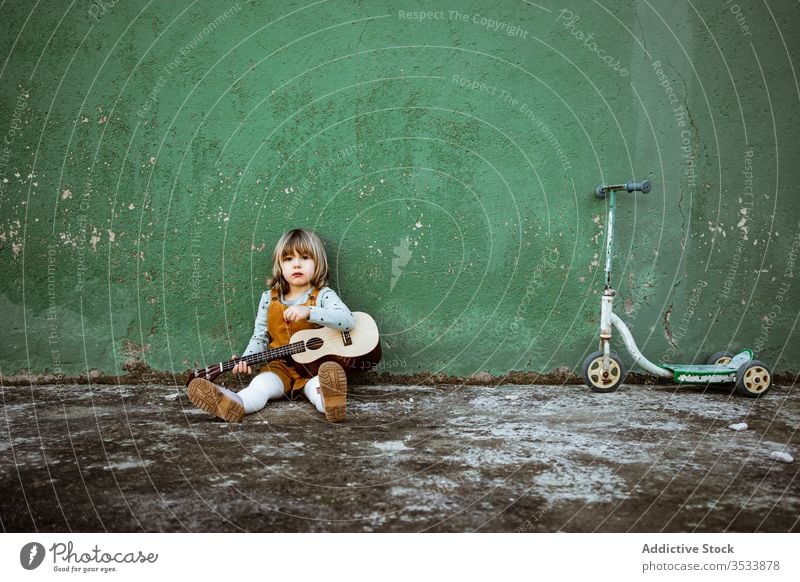 Mädchen mit Ukulele an schäbiger Wand sitzend Musik Kickroller spielen lässig wenig rau Boden verwittert grün Kind Straße Gebäude Grunge Instrument Klang Talent