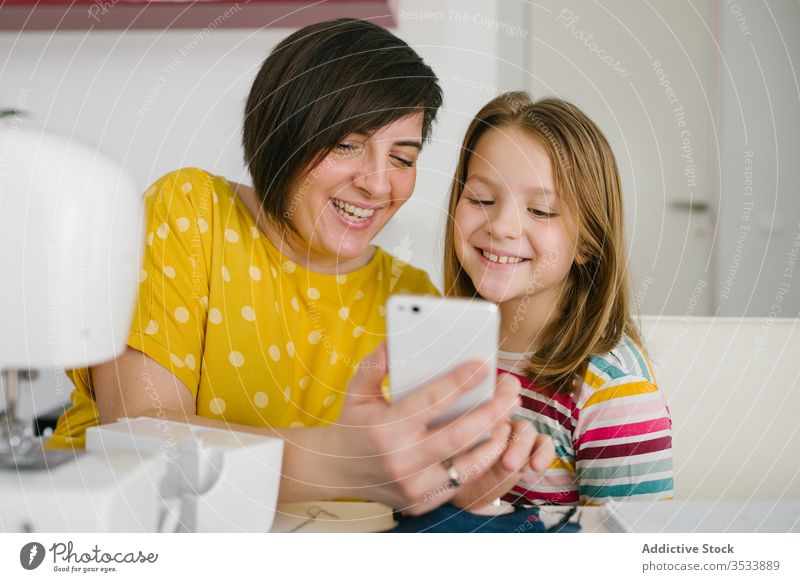 Fröhliche Näherin nimmt sich mit Tochter Mutter Selfie Smartphone Werkstatt Schneider Lächeln heimwärts Arbeit Zusammensein Frau Mädchen Tisch heiter